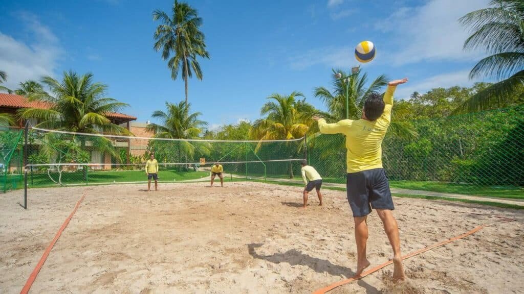 A imagem mostra um grupo de quatro homens jogando vôlei em uma quadra de areia ao ar livre, cercada por uma rede de proteção verde e vegetação tropical. Os jogadores usam camisetas amarelas e shorts, e um deles está se preparando para sacar a bola. Ao fundo, há palmeiras altas e um prédio com telhado de telha vermelha, completando o cenário ensolarado e tropical do Salinas Maragogi All Inclusive Resort, um dos melhores resorts all inclusive no Brasil.