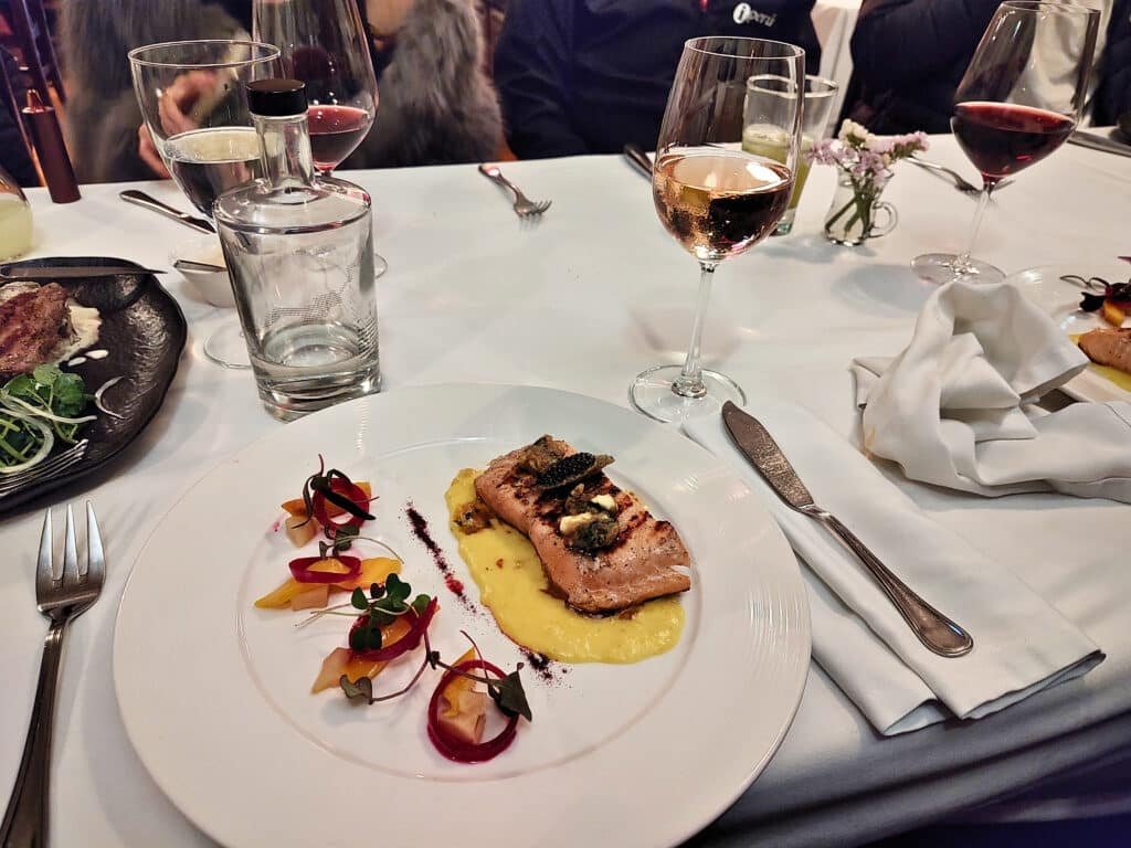 mesa com toalha branca e um prato de comida em cima, onde há um pedaço de salmão grelhado, com purê de batatas e outras especiarias. Além disso, há taças de bebidas acompanhando o prato no restaurante Cicciolina.
