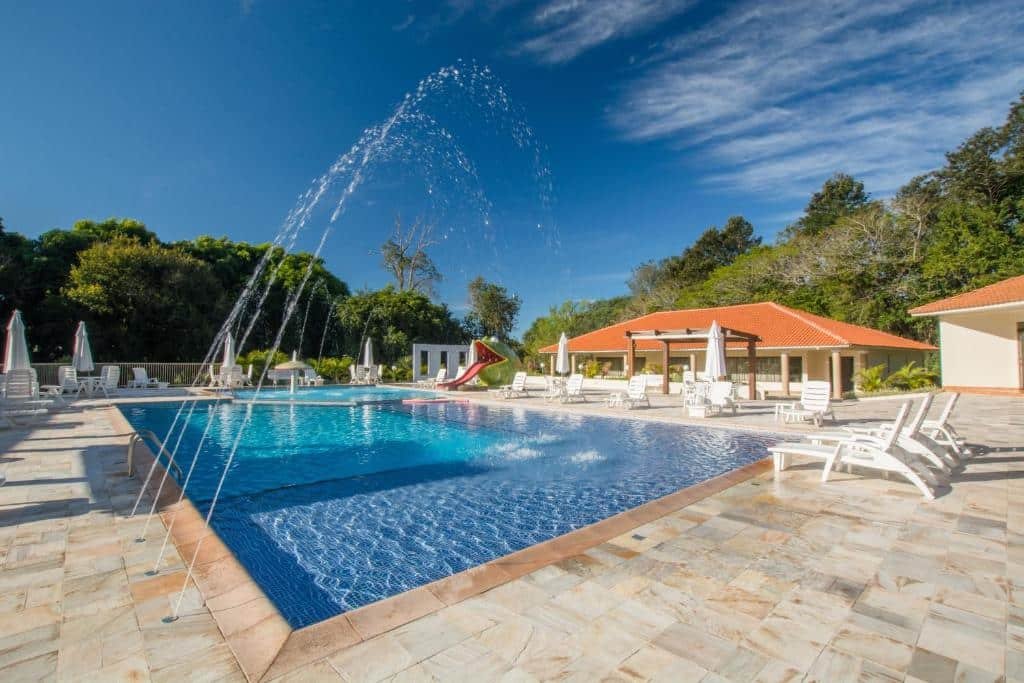Parte do Eco Cataratas Resort by San Juan que mostra uma grande piscina ao ar livre com cascatas de água. Em volta da piscina tem algumas espreguiçadeiras e mesas com guarda-sóis, algumas árvores e uma construção ao fundo. Imagem ilustrando post Resorts em Foz do Iguaçu.