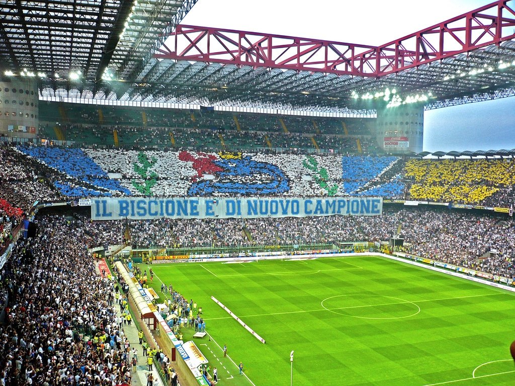 Estádio San Siro cheio de fãs exibindo uma grande faixa e mosaico suspenso em uma partida de futebol. O campo é visível em primeiro plano. A foto está na lista de o que fazer em Milão. Essa é uma opção de o que fazer em Milão para os fãs de futebol.