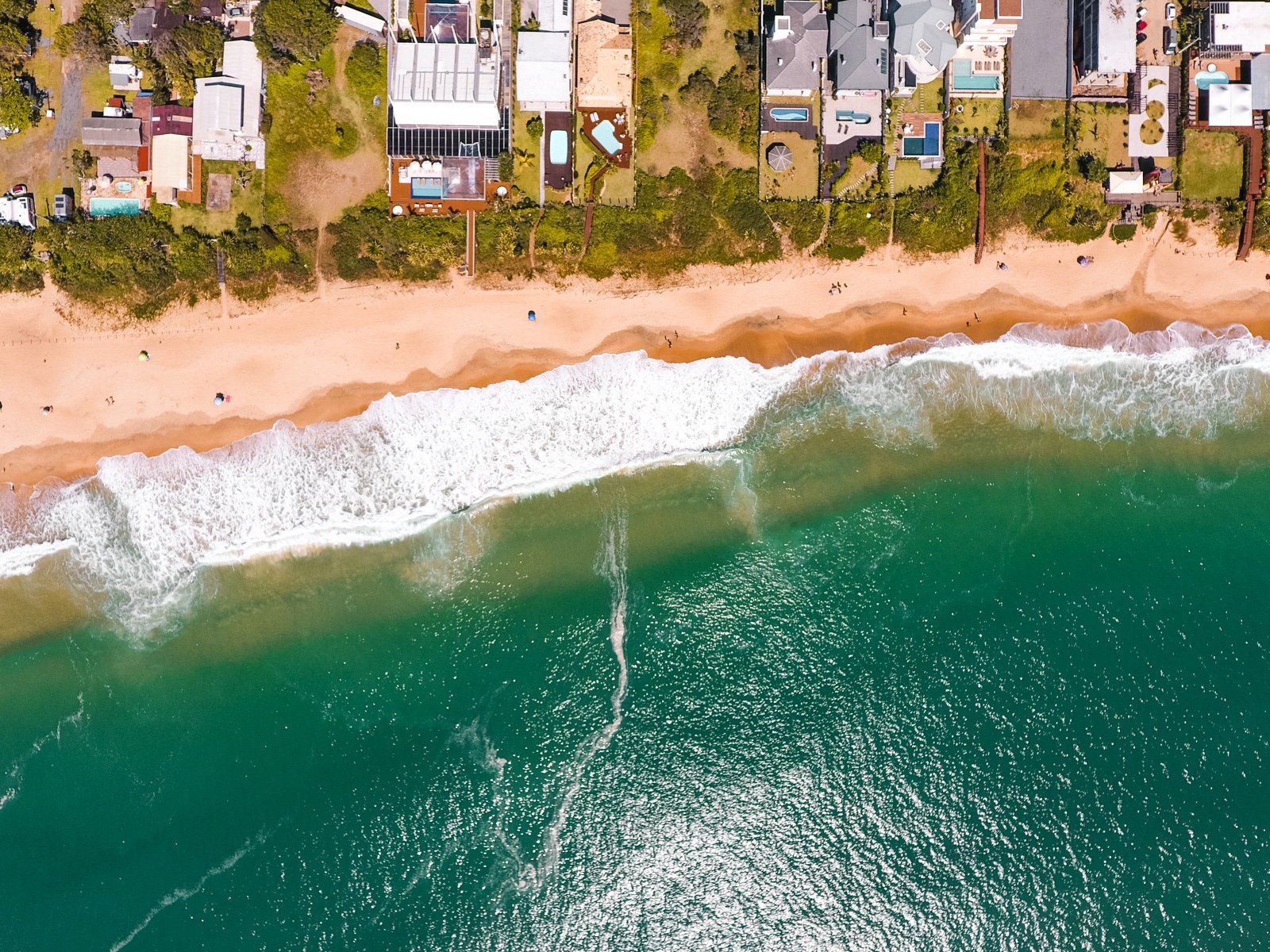 Imagem de cima da Praia do Estaleirinho. Um mar verde cristalino, a orla da praia, e no fundo propriedades.