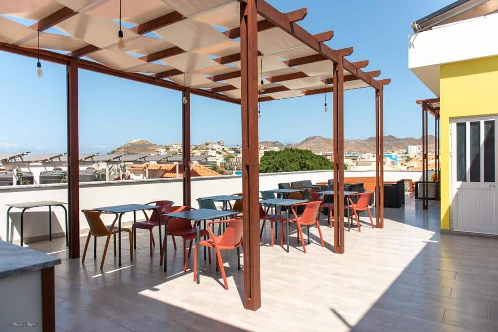 Parte do terraço Santa Cruz Boutique Hotel em Mindelo que mostra uma cobertura feita de madeira com várias mesas com cadeiras em baixo. O local tem vista para as outras construções e  montanhas ao fundo. Imagem ilustrando post Hotéis em Cabo Verde.