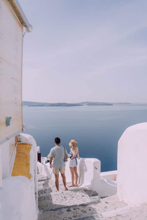 Um casal está em uma escadaria de pedra branca com vista para o mar, com ilhas distantes sob um céu claro em um cenário costeiro.