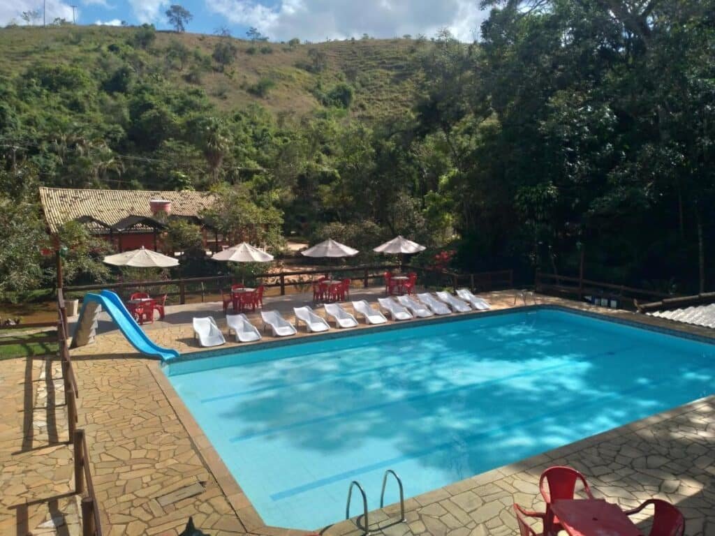 Piscina ao ar livre no Hotel Fazenda Recanto Sarandy com espreguiçadeiras brancas e um escorregador azul, cercada por árvores e colinas. Cadeiras e mesas vermelhas sob guarda-sóis nas proximidades. Esse é um dos hotéis fazenda no Rio de Janeiro.