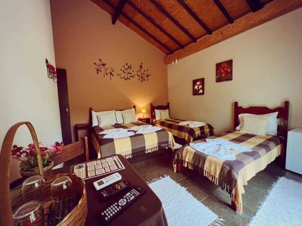 Um quarto aconchegante do Hotel Fazenda Recanto Sarandy com duas camas, cobertores xadrez, uma geladeira pequena, uma cesta e decorações de parede em um ambiente rústico.