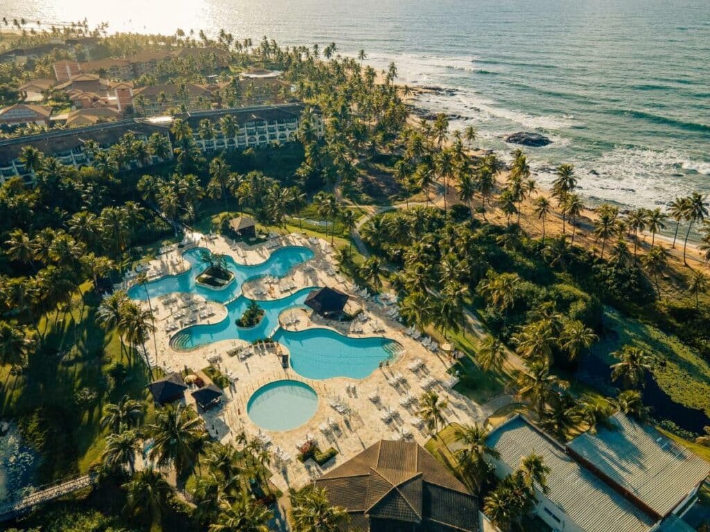 A imagem aérea mostra o complexo Sauípe Resorts, Ala Terra All Inclusive, localizado à beira-mar. O resort possui diversas piscinas com formatos orgânicos, rodeadas por espreguiçadeiras e guarda-sóis. A área das piscinas é cercada por coqueiros e outras vegetações tropicais, que proporcionam sombra e um ambiente natural. Ao fundo, pode-se ver a praia e o mar, com ondas suaves batendo na costa. As construções do resort estão integradas à paisagem, com telhados de cerâmica e arquitetura que combina com o ambiente tropical.