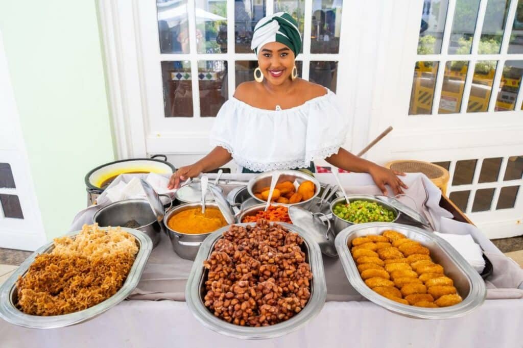 Na imagem, uma mulher sorridente, vestida com trajes tradicionais, está em frente a uma mesa cheia de comidas típicas no Sauipe Resorts Ala Terra - All Inclusive, um dos melhores resorts all inclusive no Brasil. Ela usa uma blusa branca de ombros de fora e um turbante verde e branco. A mesa está repleta de diversos pratos coloridos, incluindo feijão, farofa, acarajé e vegetais, que remetem à culinária baiana. Ao fundo, há uma porta com vidros que deixa entrever parte do ambiente externo.