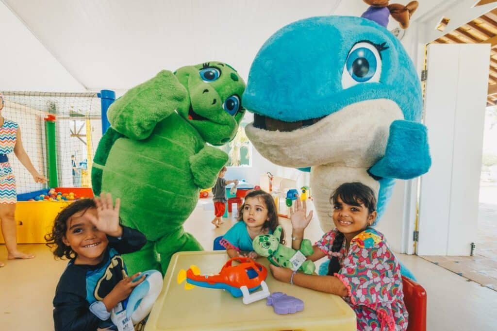 Na imagem, três crianças estão sentadas ao redor de uma mesa de brinquedos, sorrindo e acenando para a câmera no Sauipe Resorts Ala Terra - All Inclusive, um dos melhores resorts all inclusive no Brasil. Elas estão acompanhadas por duas mascotes fantasiadas: uma tartaruga verde e uma baleia azul, que estão interagindo de forma amigável com as crianças. O ambiente é claro e colorido, com uma área de recreação ao fundo, onde há uma piscina de bolinhas.