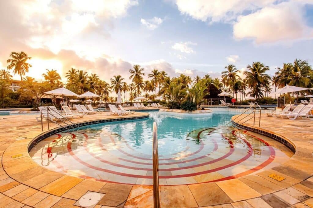 A imagem mostra uma ampla área de piscina no Sauipe Resorts Ala Terra - All Inclusive com espreguiçadeiras dispostas ao redor. A piscina tem degraus de entrada e é cercada por palmeiras. O céu está parcialmente nublado, com o sol nascendo ou se pondo ao fundo, criando um ambiente tranquilo e convidativo. Algumas áreas da piscina têm bordas arredondadas, dando um toque elegante ao espaço.