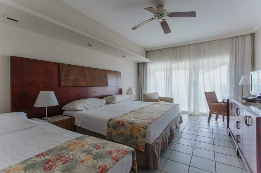 A imagem mostra um quarto do Sauipe Resorts Ala Terra - All Inclusive, um dos melhores resorts all inclusive no Brasil. Há duas camas de casal com cabeceiras de madeira escura e roupas de cama claras, decoradas com colchas florais. À direita, há uma varanda com cortinas brancas translúcidas, que permitem a entrada de luz natural. Um ventilador de teto está instalado acima das camas. No canto, há uma cadeira e uma mesa, sugerindo um ambiente confortável e relaxante.