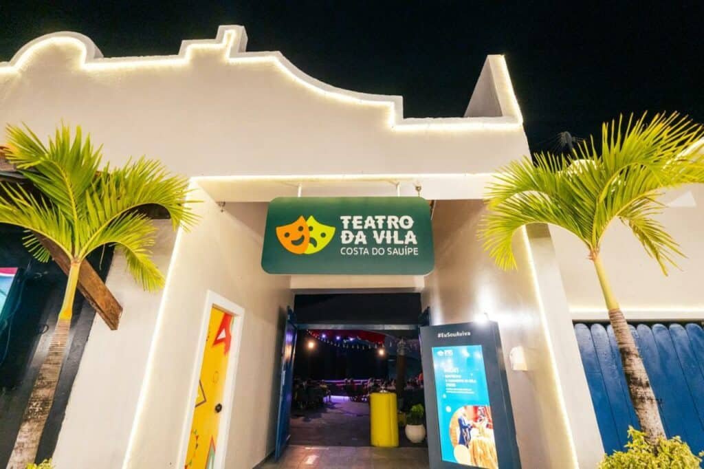 A imagem mostra a entrada do "Teatro da Vila" localizado na Costa do Sauípe. A fachada do teatro é branca, com linhas arquitetônicas simples e uma iluminação suave que destaca os contornos do prédio. À frente da entrada, há um letreiro verde com duas máscaras de teatro, uma sorridente e outra triste, acompanhadas do nome do teatro. Duas palmeiras estão posicionadas simetricamente ao lado da entrada, contribuindo para uma atmosfera tropical e convidativa. A porta está aberta, revelando o interior iluminado do teatro, onde algumas pessoas já estão sentadas.