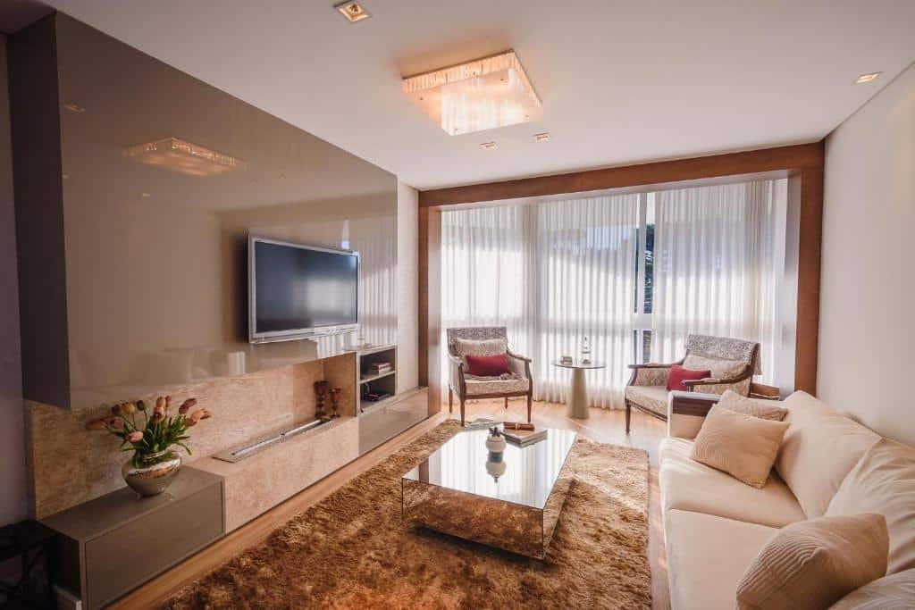 Sala no Serra Nevada Apartamento. O ambiente é espaçoso, Há um sofá à direita, uma mesa de centro à sua frente e um painel com televisão. No fundo, há duas poltronas e uma janela grande que percorre toda a parede.
