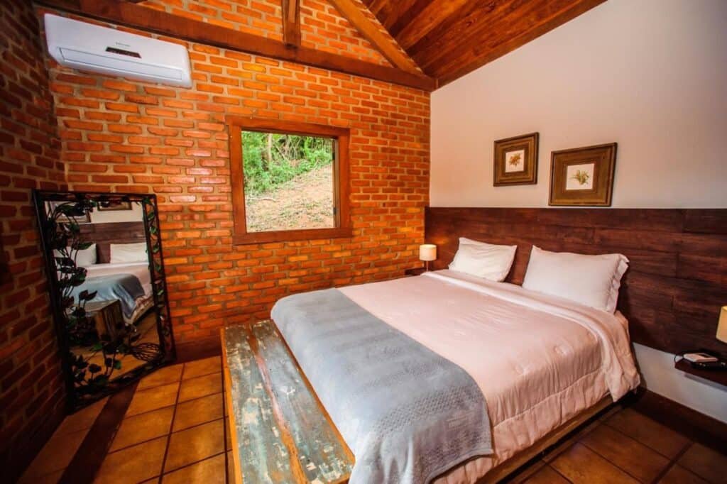 Um quarto aconchegante do Serra do Ibitipoca Hotel de Lazer com uma cama de casal, parede de tijolos, janela, ar condicionado, espelho, teto de madeira, quadros de arte e luminárias de cabeceira.