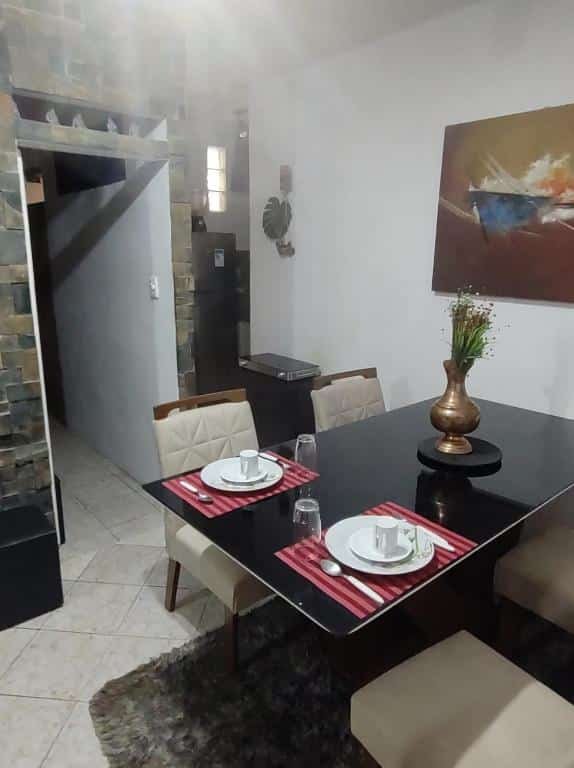 Mesa de jantar em Serra Negra - Melhor localização da cidade. É uma mesa de vidro preta, com quatro cadeiras. Há um conjunto de talheres em cima dela, e um vaso ao centro. No fundo há um corredor não iluminado.