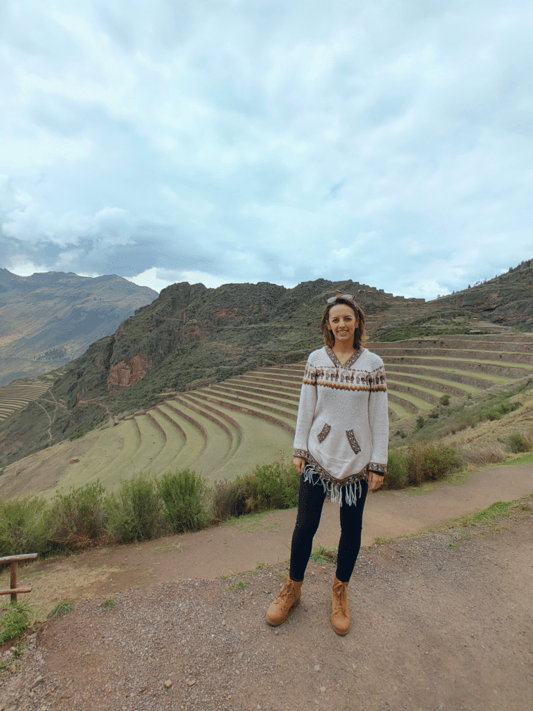 paisagem do setor agrícola do sítio arqueológico de pisaq, no Vale Sagrado dos Incas, onde há vários degraus na rocha. No lado direito da imagem há uma mulher em pé, parada e sorrindo para a câmera.