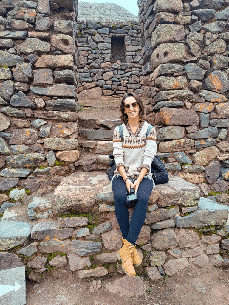 degraus e paredes de pedras empilhadas, com uma mulher sentada neles, posando para a foto. Ela está sorrindo e usa um óculos escuros marrom.