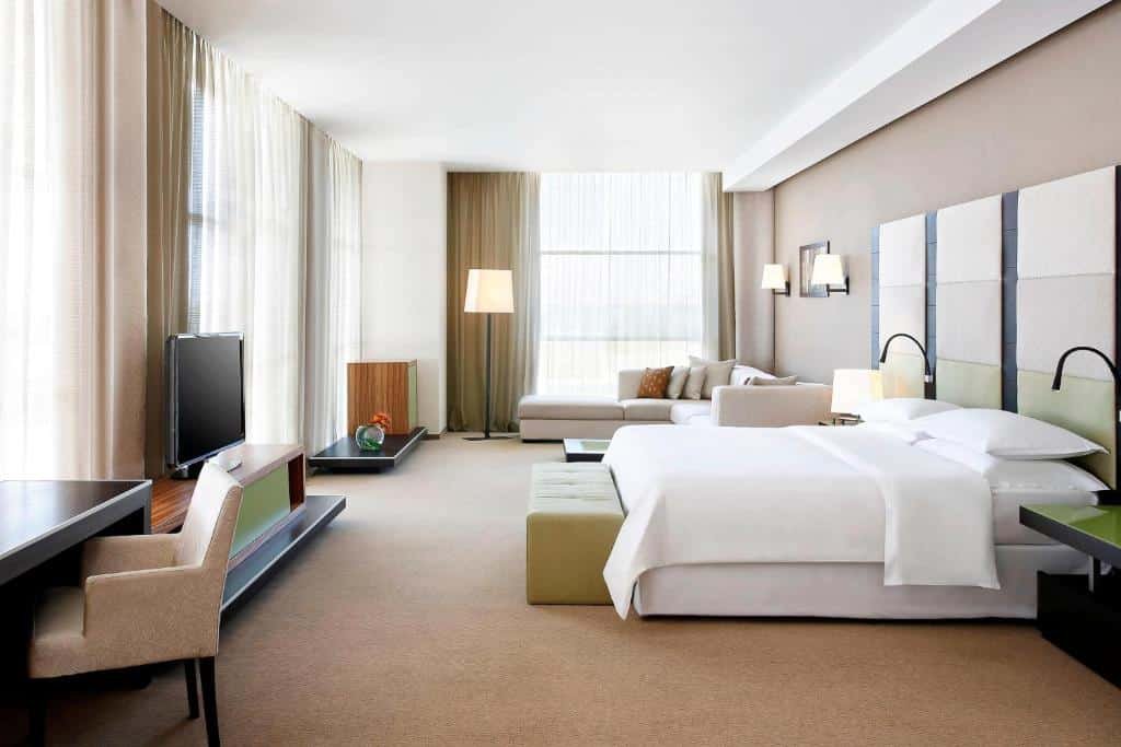 Quarto do Sheraton Milan Malpensa Airport Hotel & Conference Centre com muito espaço, há umas cama de casal, um sofá em L com almofadas, janelas amplas com cortinas, além de um móvel com uma televisão e alguns itens de decoração como abajures e vasos de plantas