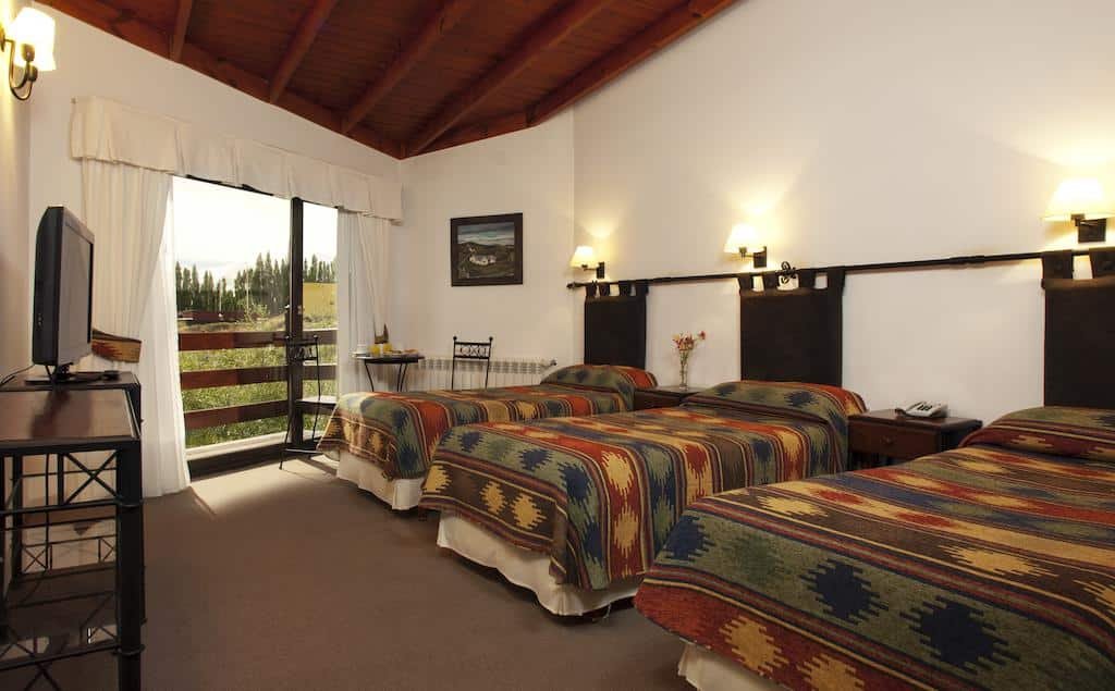 Quarto do Sierra Nevada. Três cama de solteiro do lado direito, entre elas uma cômoda. No fundo, um mesa redonda com duas cadeiras, e a janela com a sacada do quarto. De frente para as camas uma mesa com uma televisão.