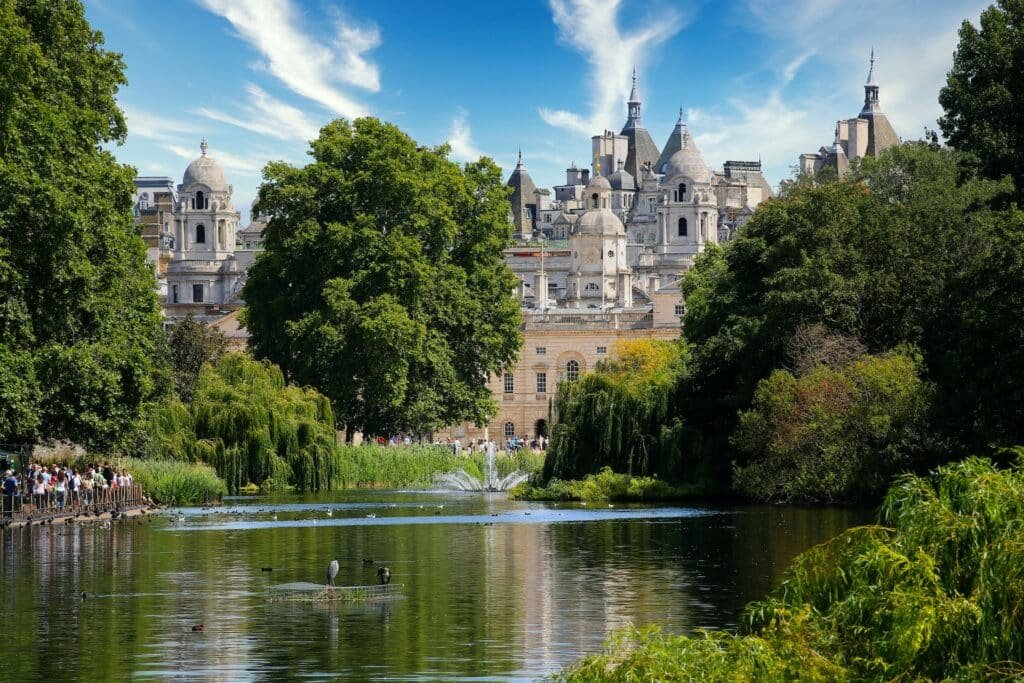 Vista do lago do Hyde Park, com muitas árvores ao redor, algumas aves nas águas e, mais ao fundo, a vista de construções que se parecem com castelos