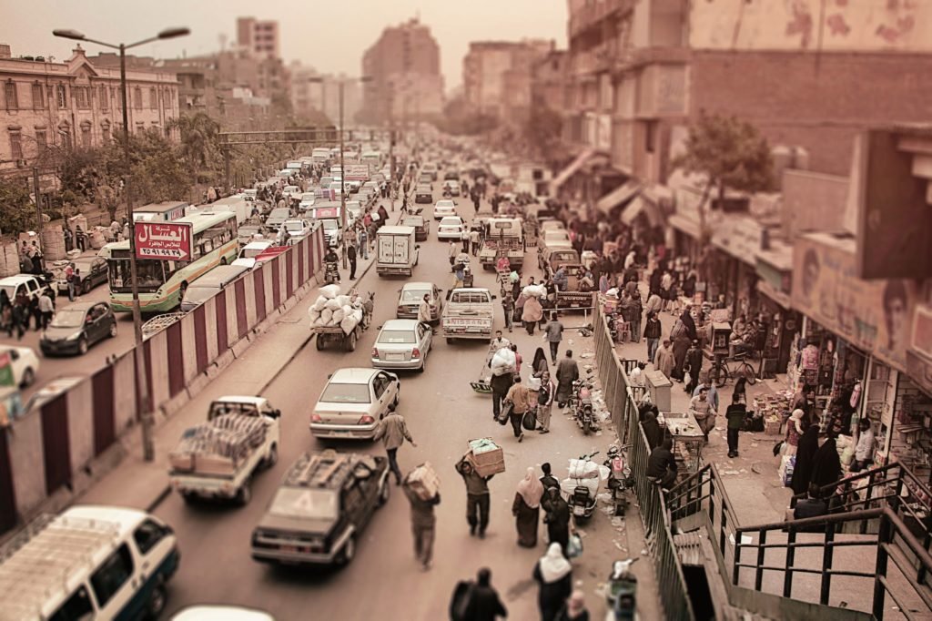 Uma rua movimentada do Cairo com tráfego pesado e pedestres. Pessoas andam pela calçada ao lado de carros e caminhões pequenos. Prédios margeiam a estrada.