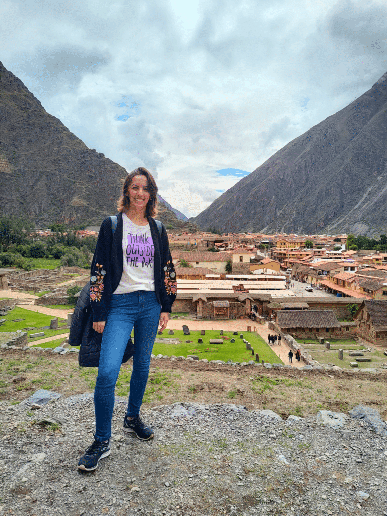 nátalie guimarães posando para foto, sorrindo e em pé, com a cidade de ollantaytambo e as montanhas peruanas do vale sagrado dos incas aparecendo ao fundo