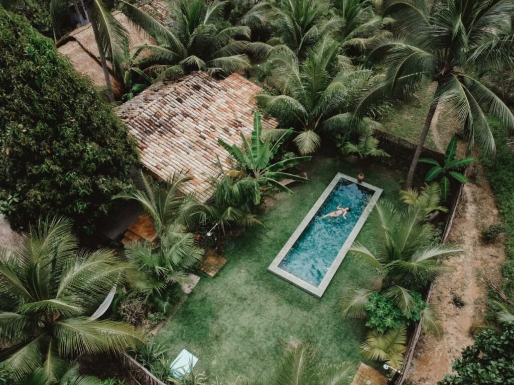 Piscina em área externa, rodeada por grama e coqueiros, com uma pessoa na água e vista do telhado de uma casa ao lado, no Sítio Peixe do Mato