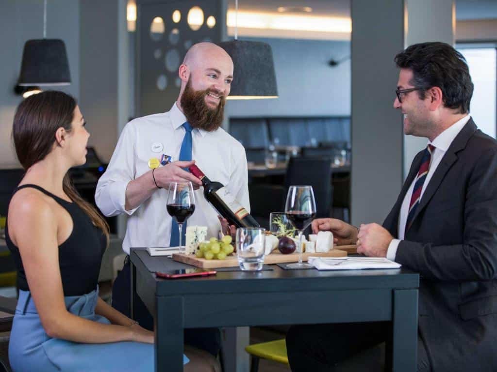 Um homem de terno sentado em uma mesa preta, de frente para uma mulher, com uma regata preta, que o acompanha. Eles prestam atenção em outro homem, um sommelier, que recomenda um vinho para eles. Na mesa, há uma tábua com queijos e frutas, copos de água e taças de vinho.