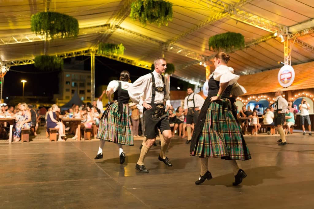 Na imagem vemos um casal dançando em primeiro plano, com trajes típicos alemães. Ao fundo há mais casais e o público que os assiste. É noite e estão em área coberta. Foto do site oficial do Parque.