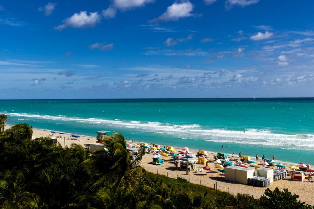 Praia de South Beach em Miami. O mar é bem azul e forma o fundo da foto. Lo em frente a ele está a faixa de areia com várias pessoas, cabanas e cadeiras. Depois, em primeiro plano, é possível ver um pedaço das árvores e gramado. A foto ilustra o post de o que fazer em Miami.