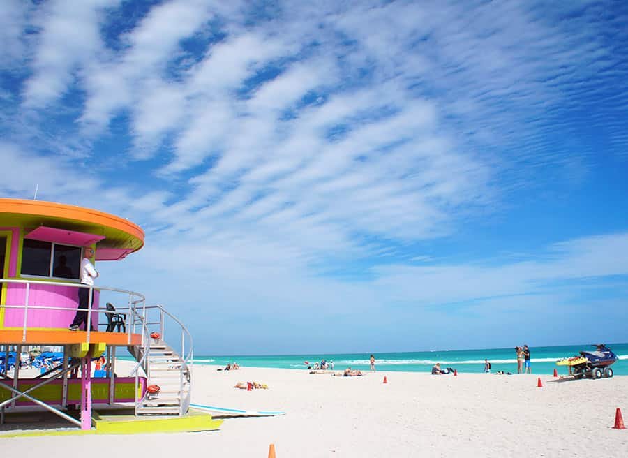 A imagem mostra a praia de South Beach, em Miami, ensolarada com areia branca e mar azul-turquesa. À esquerda, há um colorido posto de salva-vidas em tons de rosa, verde e laranja, com um guarda de pé na varanda. O céu está parcialmente nublado com nuvens finas, criando um efeito listrado. Na praia, várias pessoas estão deitadas tomando sol, nadando ou passeando. À direita, uma moto aquática está estacionada na areia. A cena é vibrante e cheia de vida, capturando um dia típico de diversão na praia.
