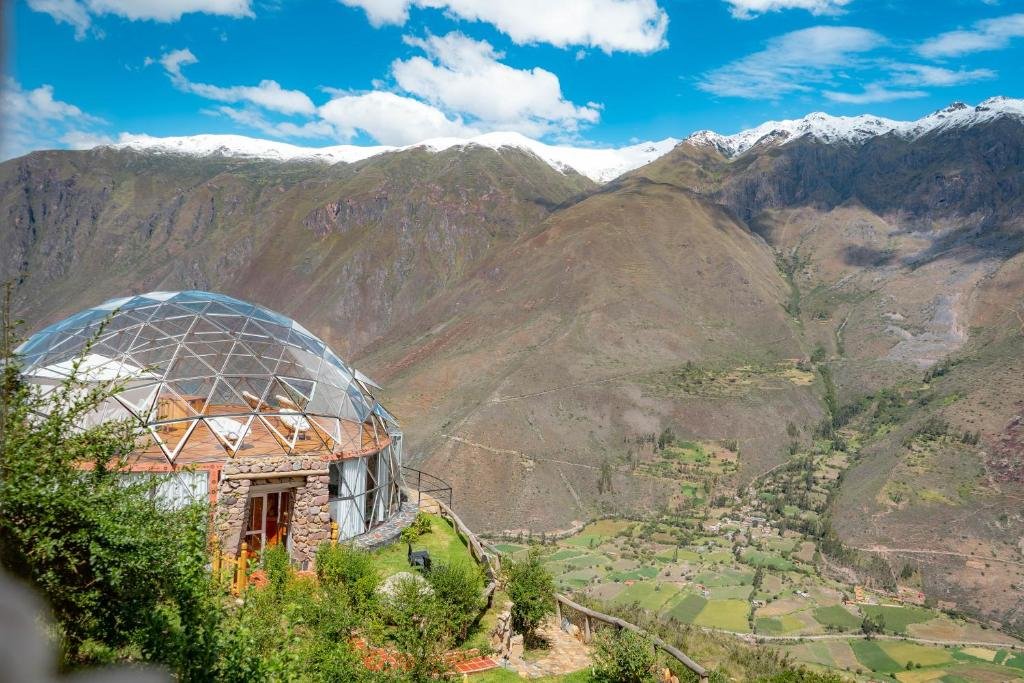 uma estrtura de metal arredondada e com teto de vidro abobado no lado esquerdo da imagem, onde fica o stardomelogde em Machupicchu, e uma cadeia de montanhas nevadas ao fundo.