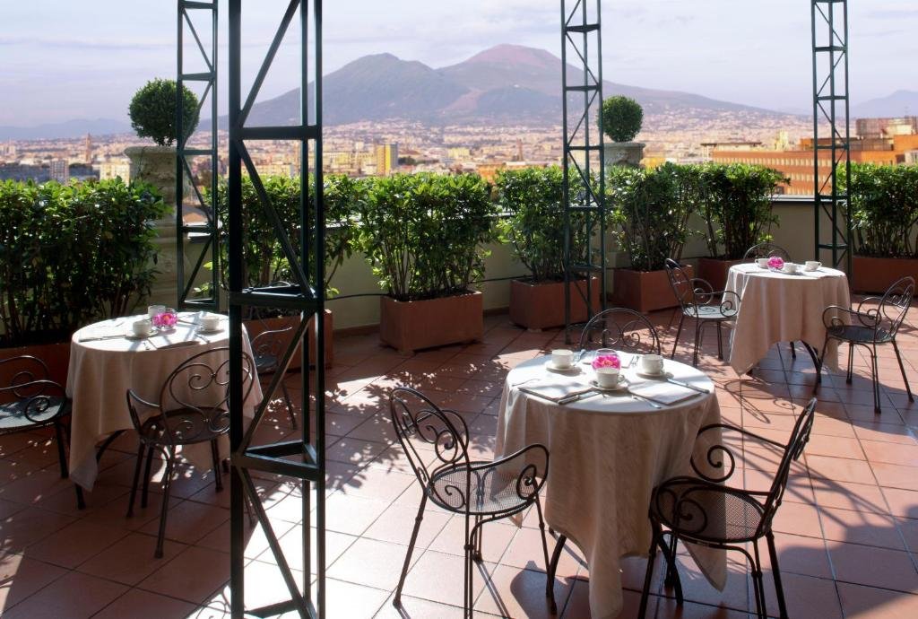 Terraço do Starhotels Terminus com três mesas redondas com cadeiras, algumas plantas verdes em volta, pilastras no meio do terraço e vista para a paisagem da cidade durante o dia, ilustrando post Hotéis em Nápoles.