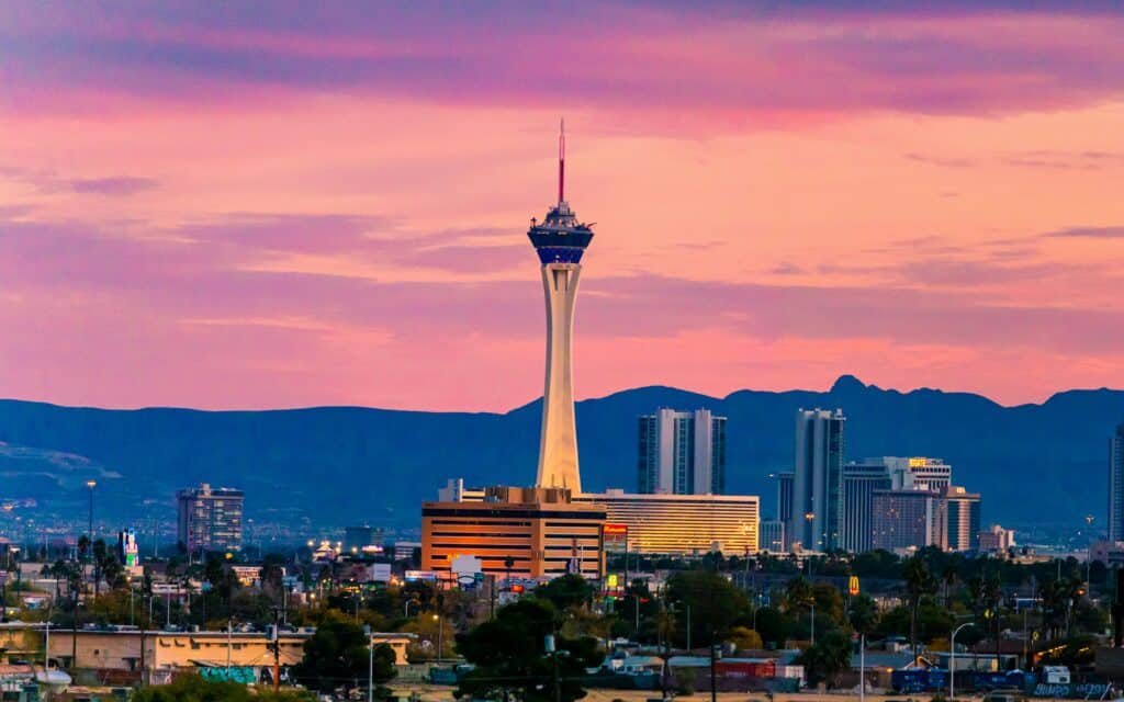Foto tirada de longe da Stratosphere Tower durante o pôr do sol. Ela é bem alta e está entre vários hotéis e árvores.