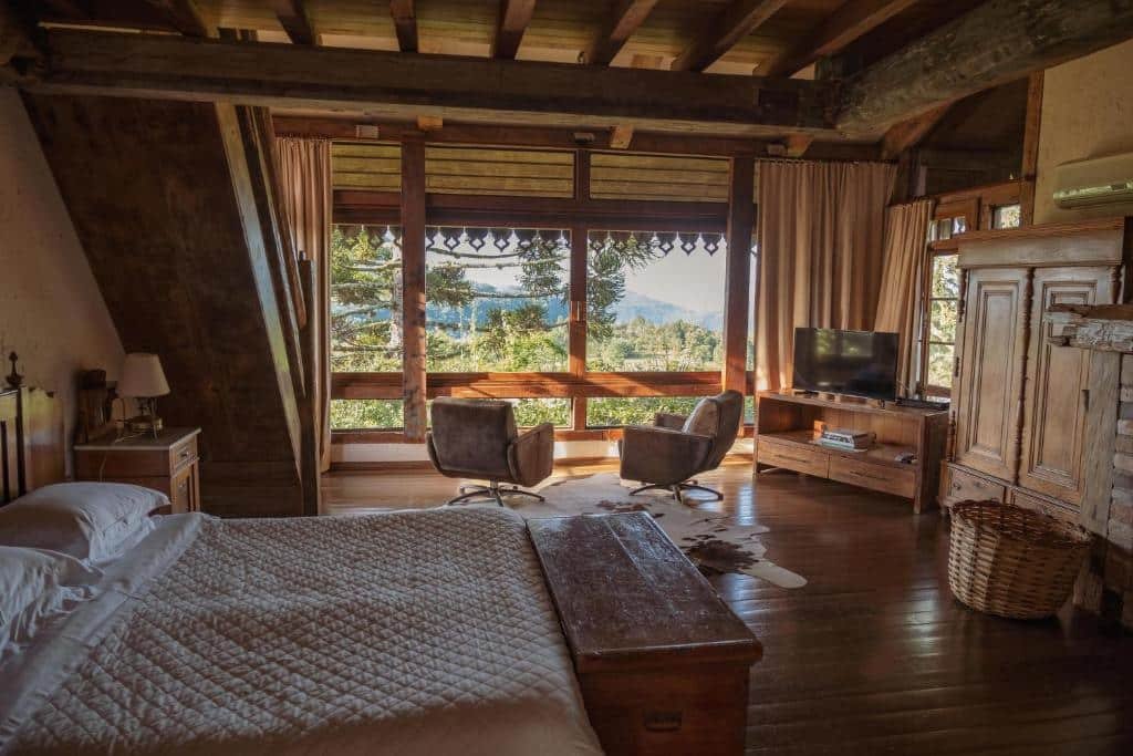 Foto do interior da Estalagem La Hacienda representando o post de chalés em Gramado. Há uma cama de casal no canto inferior esquerdo da foto Na sua frente há um armário de madeira, enquanto no fundo da foto é possível ver uma televisão e duas poltronas, além de uma grande varanda com vista para a serra.