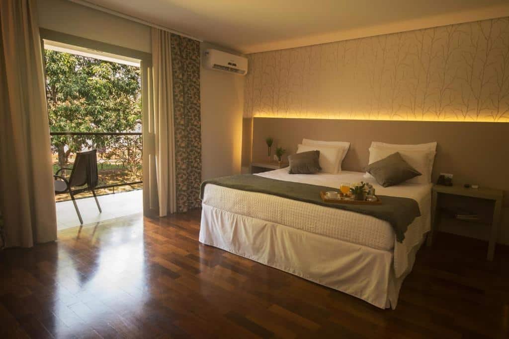 Quarto do Grand Carimã Resort & Convention Center com uma cama de casal, de cada lado da cama tem uma mesinha e um ar-condicionado do lado esquerdo. No quarto também tem uma sacada com uma cadeira e vista para as árvores.