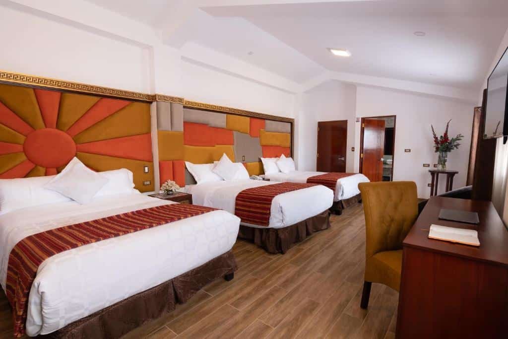 quarto familiar do Hotel Hacienda Cusco Centro Historico com três camas de casal, dispostas lado a lado, na parte esquerda da imagem. Há uma grande cabeceira estofada e colorida atrás de todas as camas. Além disso, em frente as camas há uma cômoda com mesa e uma cadeira estofada para trabalho.