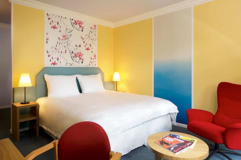 Quarto do hotel Parc Plaza. As paredes são divididas entre amarelo, azul e branco, e um papel de parede florido que está atrás da cama. A cama é de casal, e de ambos seus lados há uma pequena escrivaninha com abajur.  Na frente da cama, há uma poltrona vermelha e uma mesa redonda de centro com revistas em cima.