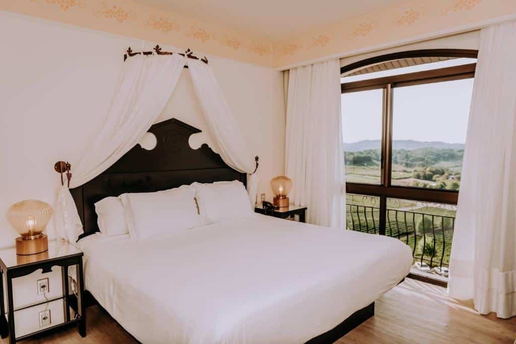 Cama de casal super espaçosa em quarto do Spa do Vinho, com mesa de cabeceira em cada lado, luminária de bronze, e uma sacada com vista para vinhedos