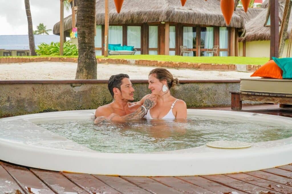 Um homem e uma mulher relaxam em uma banheira de hidromassagem ao ar livre perto de um edifício com tema tropical e telhado de palha no Summerville Resort - All Inclusive.