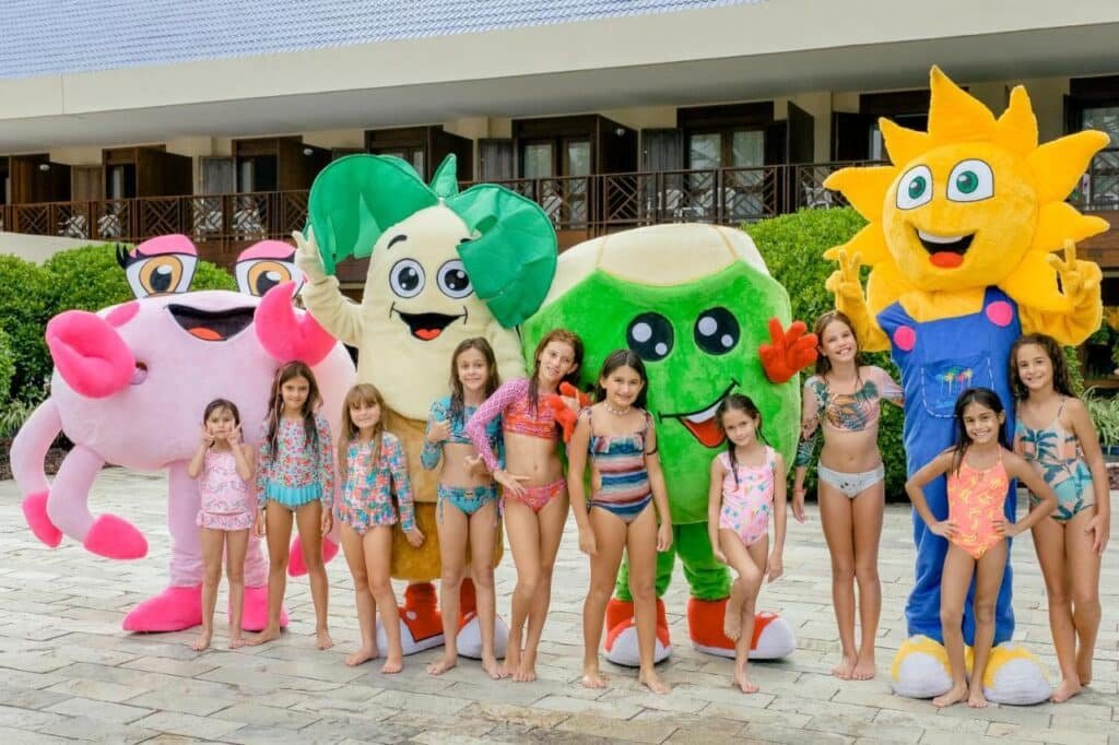 Um grupo de crianças em trajes de banho posa com grandes e coloridos mascotes de personagens de desenhos animados ao ar livre, perto do Summerville Resort - All Inclusive.