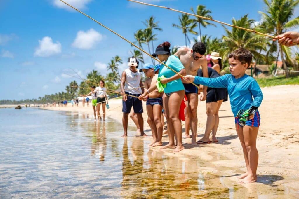 Pessoas de todas as idades estão em uma fila em uma praia ensolarada, segurando varas de pescar sobre a água. Palmeiras estão ao fundo no Summerville Resort - All Inclusive.