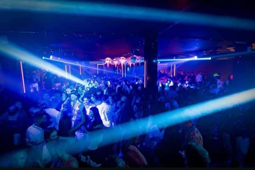 Uma boate lotada com luzes azuis e roxas iluminando pessoas dançando em um ambiente mal iluminado. Essa é a Swag Ibiza Club, uma das festas em Ibiza.