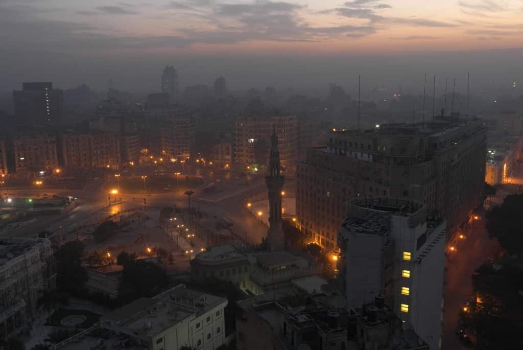 Vista aérea da praça da praça Tahrir ao anoitecer com postes de luz iluminando prédios e estátuas. O céu está cinza com nuvens pesadas. Imagem para ilustrar post sobre o que fazer no Cairo.