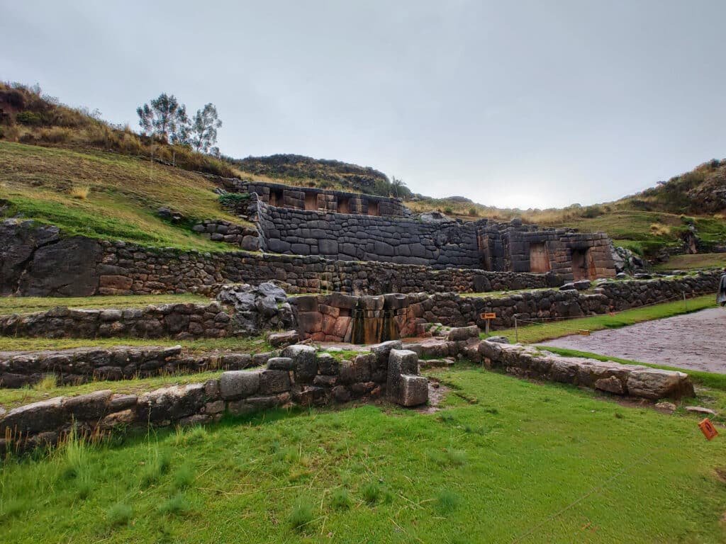 Foto do templo de pedras de granito na rocha, com vários níveis e desníveis de degraus, no Sítio Arqueológico Tambomachay, em Cusco.