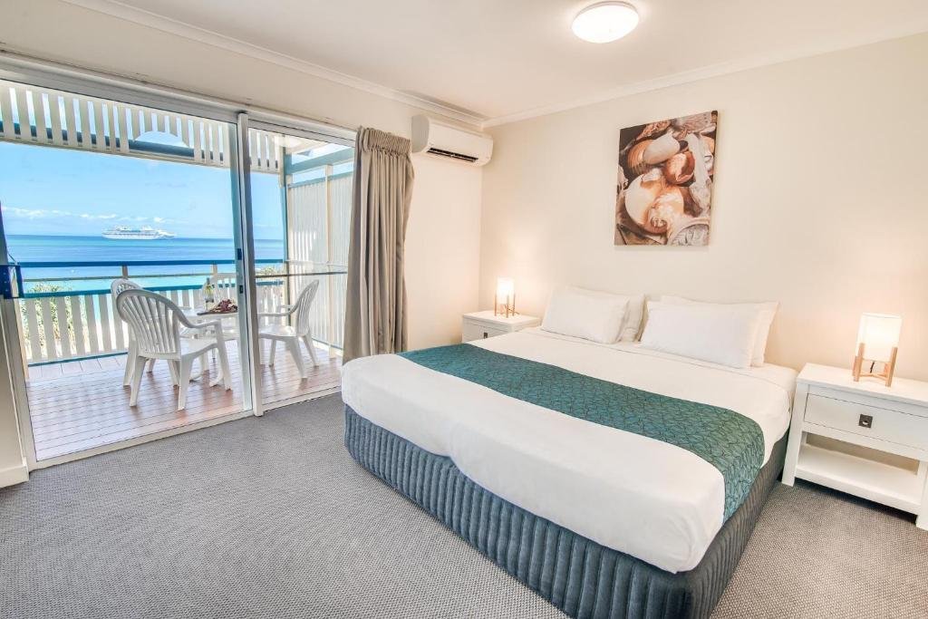 Quarto do Tangalooma Island Resort com cama de casal, roupa de cama branca e azul-petróleo, mesas de cabeceira e varanda com vista para o mar e móveis de jardim.