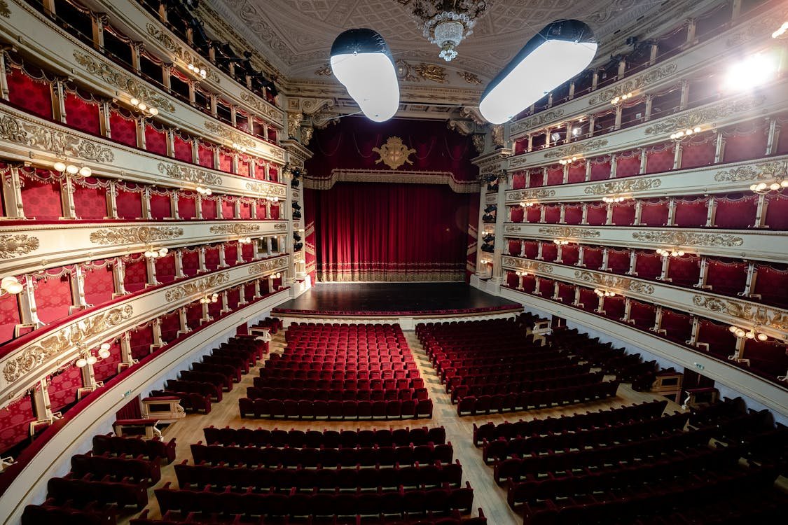 Vista do interior do Teatro alla Scala ornamentado com assentos vermelhos, sacadas, decorações complexas e um palco com uma cortina vermelha fechada. Foto ilustrando o post de o que fazer em Milão.