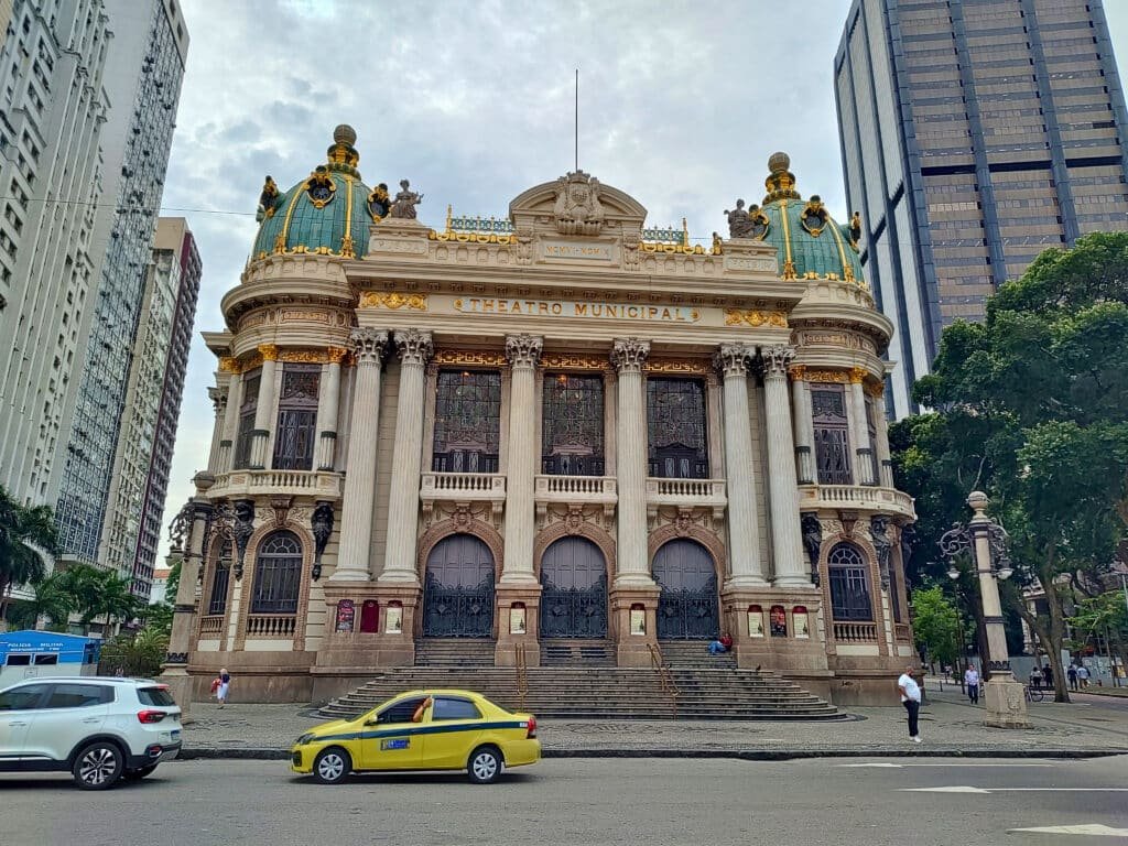 Fachada do Theatro Municipal, um edifício imponente com arquitetura neoclássica e detalhes dourados, localizado em uma área urbana. As colunas majestosas sustentam a estrutura, e as cúpulas verdes com adornos dourados adicionam um toque de elegância. Na frente, escadas levam até a entrada principal, enquanto carros e pedestres movimentam-se na rua em frente.