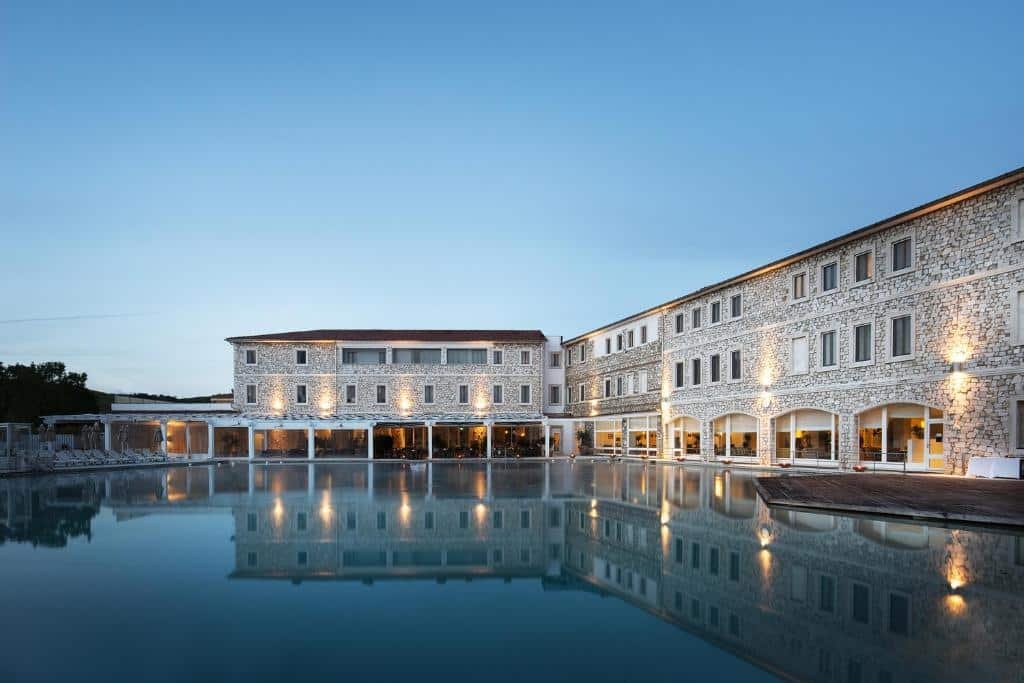 Piscina grande de um dos hotéis em Toscana, espelhando os prédios de tijos a vista do Terme di Saturnia Natural Spa & Golf Resort