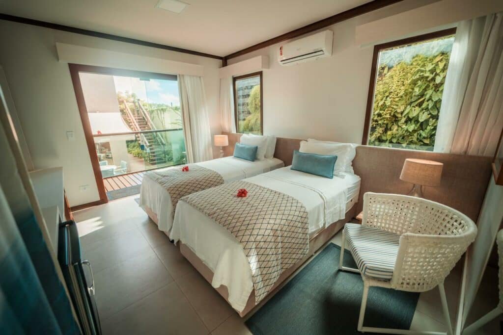 Quarto duplo da Tesouro Noronha, com duas camas de solteiro próximas, enxoval branco, porém peseira cor nude e uma almofada verde água em cada cama, janelas amplas de cada lado e ar-condicionado