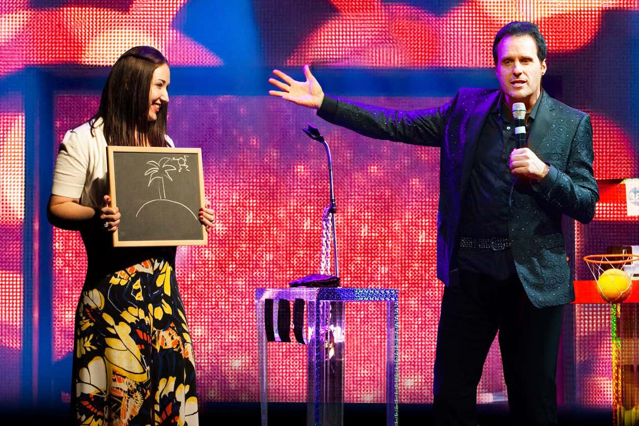Gerry McCambridge durante o show The Mentalist segurando um microfone gesticula em direção a uma mulher segurando um quadro negro com o desenho de uma palmeira, em frente a um fundo colorido.